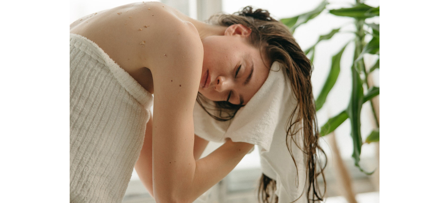 A woman in a white towel leans over with her hair wrapped in another towel, eyes closed, in a serene, spa-like setting.